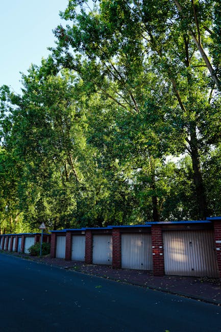 A row of closed garage doors constructed from white panels with red brick columns between each, situated along a quiet paved street with no vehicles visible. Behind the garages, there is a dense canopy of tall, leafy green trees with sunlight filtering through the foliage, creating dappled shadows on the garages and pavement. The scene depicts an outdoor setting that could be part of a residential area near Orleans Park estate, where house removals and furniture transport involve navigating narrow access points and outdoor environments. The image shows no active moving activity but highlights the typical environment where loading bays or garage access points are utilized during home relocation or packing and moving services provided by Man with Van St Margarets, which may involve using this space for loading or unloading belongings for a house move.