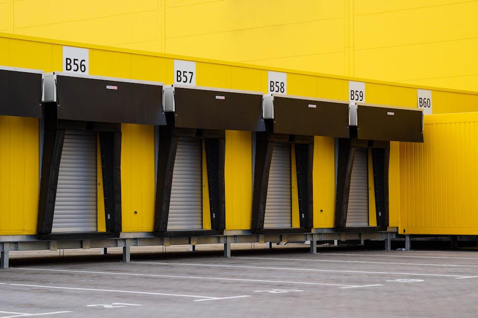 A row of four loading bays with black roll-up shutters, numbered B56 through B60, set into a large yellow industrial building. The bays are supported by black metal brackets and are situated at ground level with a paved area in front, marked with parking lines. The scene is well-lit with natural daylight, and the bays are empty, with no vehicles or equipment visible. The background shows the continuation of the yellow building's exterior wall. This setting illustrates a typical loading and unloading zone suited to home relocation and furniture transport services provided by Man with Van St Margarets.
