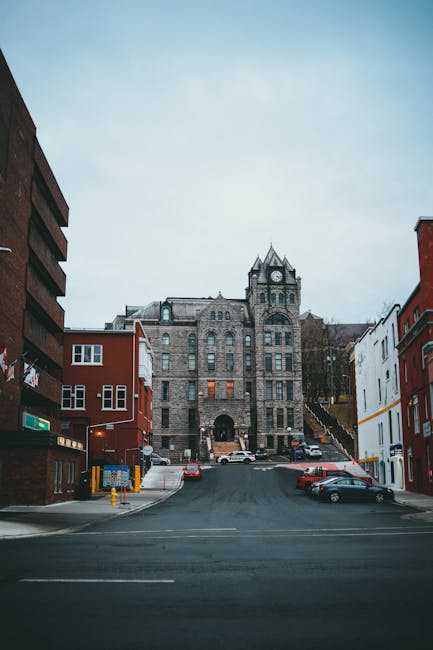 A street view taken from the pavement showing a large, historic stone building with a clock tower and arched entrance at the top of a gentle incline on St Margarets Road, TW1. On each side of the street, there are multi-storey red brick and white-painted residential buildings with modern windows, some featuring small balconies or commercial signage. Parked cars line both sides of the road, and a few vehicles are visible further up the street near the building. The street appears to be part of a residential or mixed-use area, with ambient daylight creating natural lighting. The scene reflects a typical urban environment where home relocation services, such as packing, furniture transport, and loading processes, could take place for a move scheduled on St Margarets Road. Man with Van St Margarets occasionally supports local home removals and moving logistics within this area.