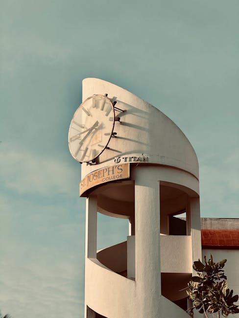 A tall, white clock tower featuring a large round clock face with black hour and minute hands, mounted high on the structure. The tower has a curved, modern architectural design with open sections supported by vertical columns, and a sign reading 'St. Joseph's College' attached to the lower part of the tower. The clock shows the time as approximately 4:15. In the foreground, a small leafy plant is visible at the base of the tower, and the background sky is partly cloudy with a soft, muted light. This scene is unrelated to house removals but captures an external view of a college building's clock tower.