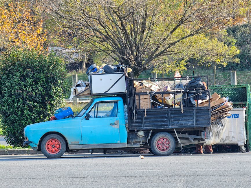 Side view of an old blue flatbed truck loaded with various household items, including large cardboard boxes, wrapped furniture, plastic containers, bags, and rolled-up carpets, parked along a suburban street during daylight. The truck's cargo area is filled with packed belongings, some secured with ropes and straps, ready for a house removal or furniture transport service. The background features a tree with green and yellow leaves, indicating an autumn setting, and a fence enclosing a garden or park area. The image reflects a typical loading process during home relocation, with the vehicle positioned on the pavement close to a residential property, demonstrating the logistics involved in packing and moving household possessions, as seen by Man with Van St Margarets.