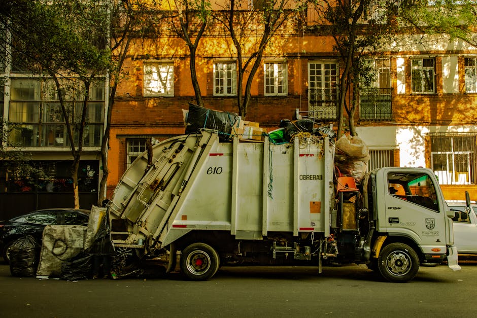 A white waste collection truck with the number 610 and 'Oberai' branding is parked on a street in St Margarets, with a height-adjustable lifting mechanism at the rear containing various recycling and waste materials, including black and transparent plastic bags, cardboard boxes, and other debris. The truck is positioned adjacent to a residential building with a brick façade, large multi-pane windows, and two trees casting shadows on the building's exterior. The scene suggests a home relocation or clearance process involving the loading or disposal of bulky items, with the truck ready for collection or transport, and nearby parked cars along the curb. Man with Van St Margarets specialises in removals and clearance services, supporting efficient household moves and waste disposal, as evidenced by the organized loading environment and equipment visible in the image.