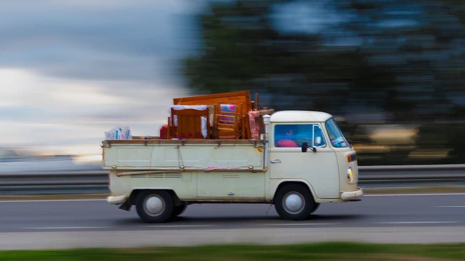 Side view of an old blue flatbed truck loaded with various household items, including large cardboard boxes, wrapped furniture, plastic containers, bags, and rolled-up carpets, parked along a suburban street during daylight. The truck's cargo area is filled with packed belongings, some secured with ropes and straps, ready for a house removal or furniture transport service. The background features a tree with green and yellow leaves, indicating an autumn setting, and a fence enclosing a garden or park area. The image reflects a typical loading process during home relocation, with the vehicle positioned on the pavement close to a residential property, demonstrating the logistics involved in packing and moving household possessions, as seen by Man with Van St Margarets.