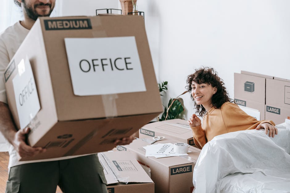 A woman with curly brown hair, dressed in a light brown top, is seated on a bed surrounded by packed cardboard boxes labeled with sizes like 'Medium' and 'Large,' and some marked 'Office.' She is smil
