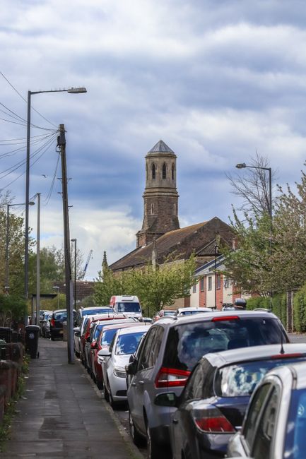 A street scene showing a row of parked cars lining a pavement on one side of the road, with a variety of vehicles including hatchbacks and SUVs, some with roof racks, all parked close together. The pavement is narrow and bordered by residential houses with gardens, trees, and bushes, and a tall, historic stone church with a square tower and a pointed roof is visible in the background under a cloudy sky. Utility poles with overhead cables run parallel to the street, while streetlights are mounted on poles along the pavement. The scene captures an everyday setting in St Margarets, with vehicles possibly awaiting loading or unloading as part of a house removal or moving process, coordinated by local movers like Man with Van St Margarets.
