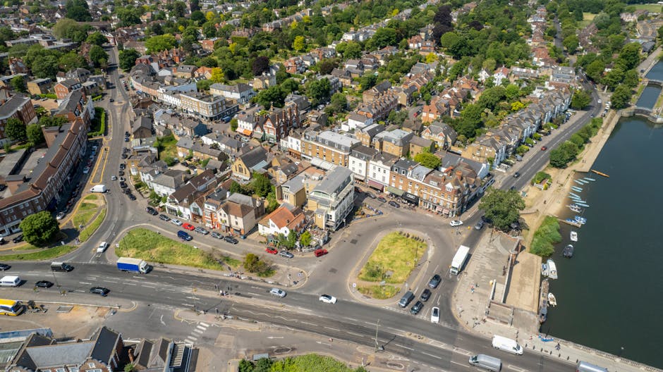 An aerial view of a residential area in St Margarets, Richmond upon Thames, showing a mix of terraced houses, apartment buildings, and shops along the main street. The scene includes a small parking area with several cars and a few delivery vans parked near the buildings. To the right, there is a river with boats moored along the bank, adjacent to a paved walkway. In the foreground, a road with multiple lanes contains moving vehicles, including trucks and cars, some of which are part of a home relocation process. The area is shaded by numerous trees and surrounded by lush greenery, with well-maintained pavements and urban infrastructure visible, all captured during daylight hours. This setting exemplifies typical urban logistics involved in furniture transport, packing, and loading operations associated with professional removals, such as those offered by Man with Van St Margarets.