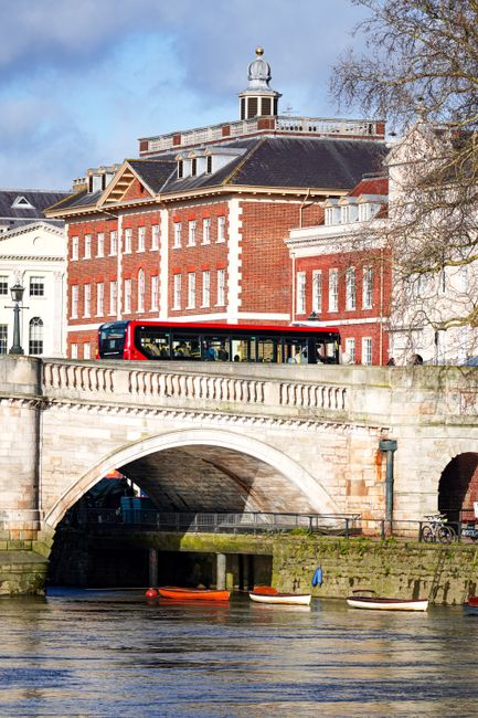 A red and black bus is visible in the foreground, parked alongside a stone bridge with ornate balustrades, which spans over a river with small boats moored along the bank. Behind the bridge, there is a large red brick building with multiple white-framed windows, a dark slate roof, and a prominent clock tower at the top. The scene is set outdoors in daylight, with a clear sky and some trees with bare branches on the right side. The image captures a part of the home relocation and moving process involving transportation logistics, with the building and bridge hinting at the typical urban environment of Richmond upon Thames. Man with Van St Margarets, a professional removals company, would handle such a move, often involving packing, loading furniture and boxes, and transporting goods via nearby roads and bridges, similar to the scene depicted.