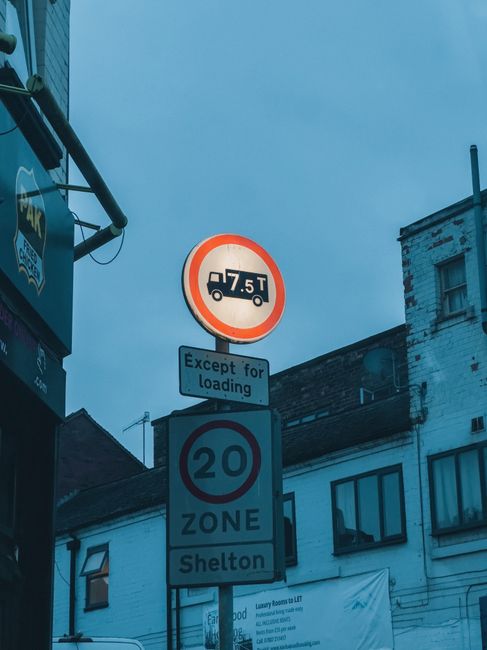 A street scene in St Margarets showing a vertical street signpost with multiple signs attached, including a red and white circular sign indicating a maximum vehicle weight of 7.5 tonnes, with an additional smaller sign below it stating 'Except for loading.' Below these, a blue circular sign displays the speed limit of 20 miles per hour and indicates a 'ZONE' for Shelton. The background features a blue sky and a mix of residential buildings, some with white and brick facades, and windows. A traffic-light pole, partially visible, is on the left side of the image, along with a green overhead street arm. This scene is relevant to house removals and packing and moving services, such as those offered by Man with Van St Margarets, especially when adhering to local street loading regulations during home relocation or furniture transport processes.