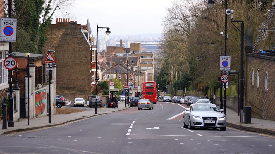 A street scene in St. Margarets showing a residential area with a mix of brick houses and trees lining the pavement. In the foreground, a grey Audi vehicle is parked on the right side of the road, while a white sedan is approaching from behind. On the left, a small group of people is gathered near a bus stop, adjacent to a red London double-decker bus that is further down the street, heading away from the camera. The road has a gentle curve and is marked with street lines and directional arrows. Several parking spaces along the curb are occupied by various cars, and there are traffic signs including a 20 mph speed limit and controlled zone signs indicating parking restrictions. The environment appears well-lit with natural daylight, and streetlights and lampposts run along both sides of the street, providing a typical urban setting suitable for home relocation or furniture transport activities, as managed by Man with Van St Margarets.