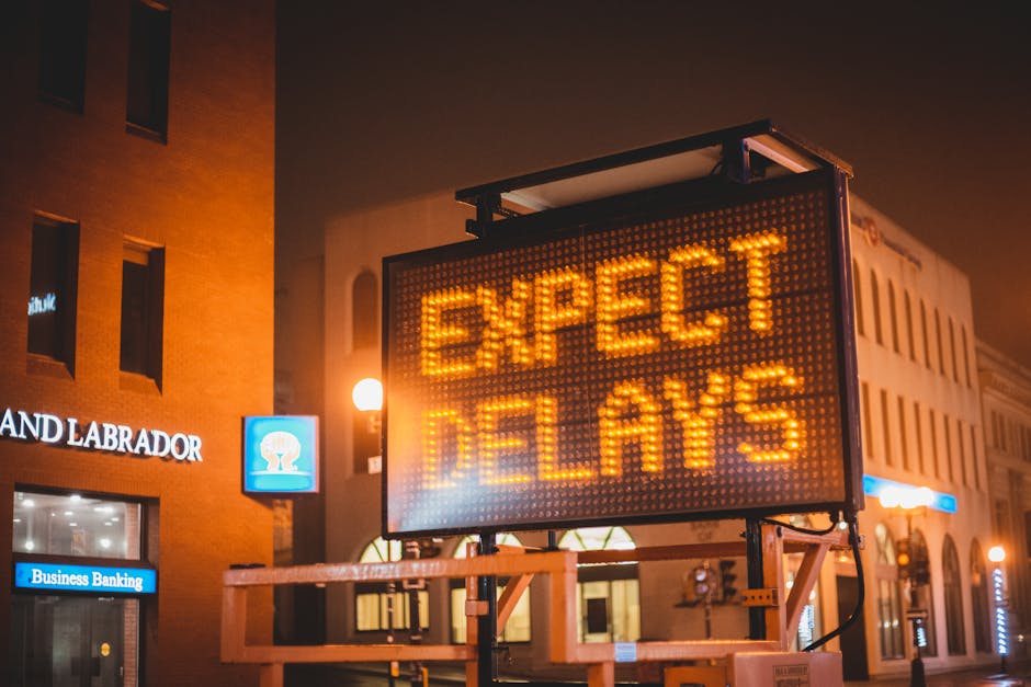 A brightly illuminated digital road sign displaying the message 'EXPECT DELAYS' in orange LED lights is mounted on a metal frame with a wooden platform at the base, situated outdoors on a city street 
