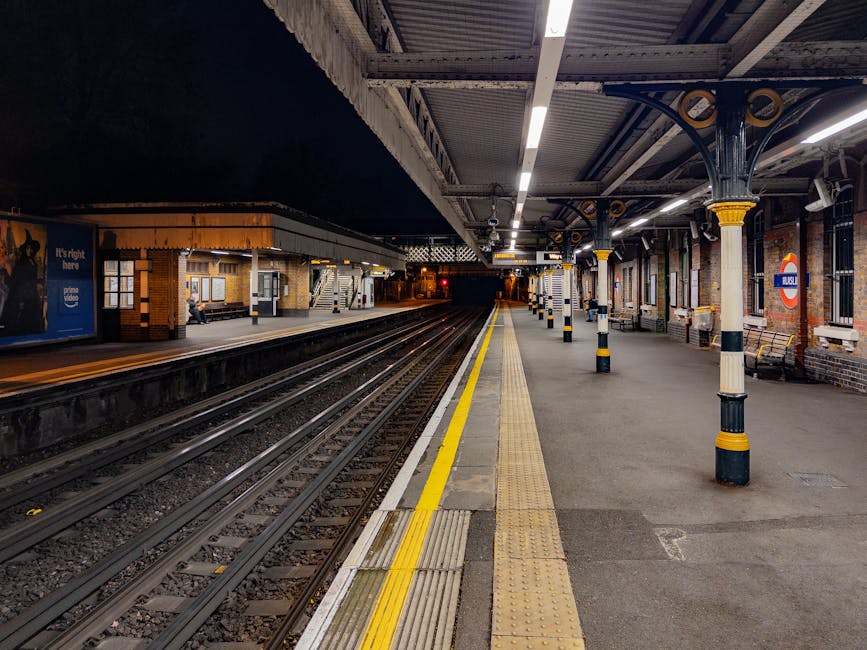 A vacant platform at St Margarets railway station during nighttime, featuring two parallel train lines with dark rails and wooden sleepers, bordered by a yellow safety line along the edge. The platform surface is made of grey concrete with tactile paving strips near the edge for safety. The station is sheltered by a metal roof supported by black and yellow decorative cast iron columns, with lighting fixtures casting a bright white light across the area. On the far side of the platform, brick station buildings with windows and seating are visible, one with a person sitting near the entrance. An illuminated ‘London Underground’ roundel sign is mounted on the brick wall near the platform’s edge. The setting appears quiet, with no trains present and minimal activity, reflecting a scene suitable for a home relocation process involving the careful transport of belongings via nearby transport services, as managed by companies like Man with Van St Margarets.