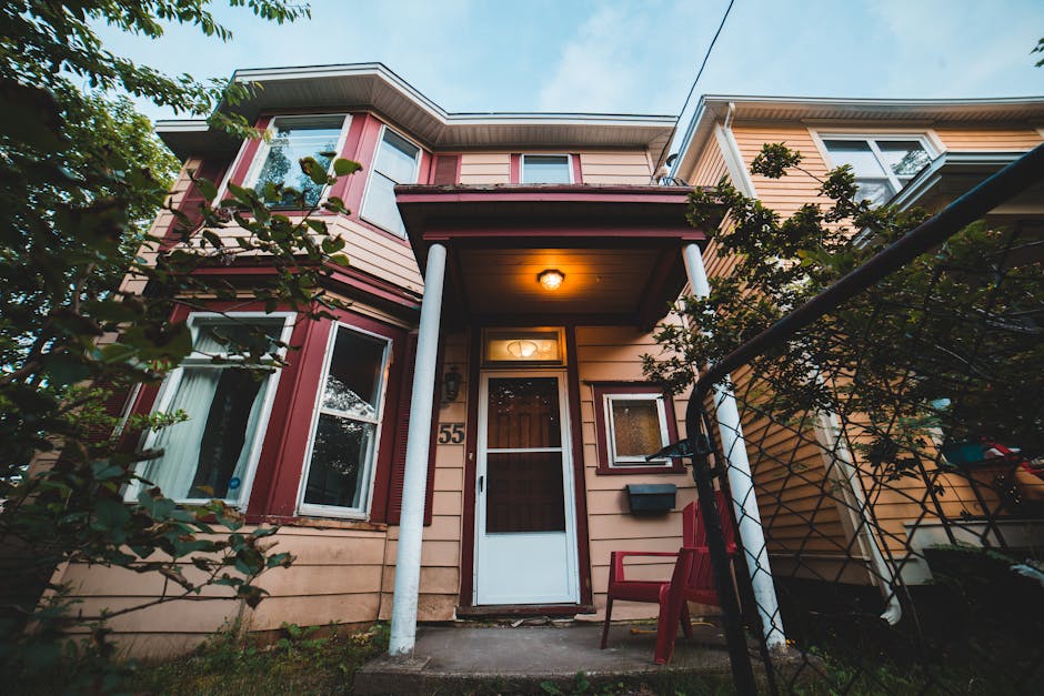 The image depicts the exterior of a multi-story residential house with beige and reddish trim, featuring several windows and a white front door with a glass panel. The house has a small porch area with a roof supported by two white pillars, illuminated by a warm exterior light. There is a red plastic chair positioned near the entrance, and the house number '55' is visible beside the door. In the foreground, a black metal fence partially surrounds the property, with some green foliage from nearby plants and bushes on the left side of the image. The setting appears to be during twilight, with natural daylight fading and the porch light providing additional illumination. This scene is relevant for house removals, as it shows a typical residential property where furniture and packing materials could be prepared for a home relocation or moving process, with emphasis on the exterior environment, entryway, and surrounding area often involved in the logistics handled by Man with Van St Margarets.