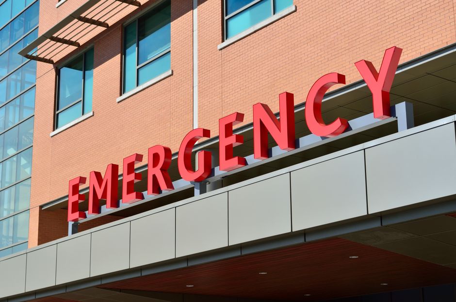 Exterior view of a modern building with a large red 3D sign reading 'EMERGENCY' mounted above the entrance. The building features a brick facade and multiple windows, with part of a glass-walled staircase visible on the left side. The entrance area has a gray overhang with a wooden underside, possibly serving as a loading zone for emergency or urgent move services. The scene is well-lit with natural daylight. This setting relates to urgent eviction moves and home relocation services provided by Man with Van St Margarets, who specialize in removals and furniture transport, including loading and packing processes.